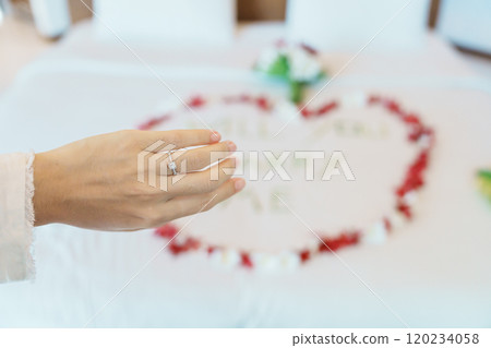 Woman hand wearing a diamond ring after say YES with message WILL YOU MARRY ME made of flower petals with bouquet on bed. Idea for marriage proposal, romantic surprise, wedding and engagement concept 120234058
