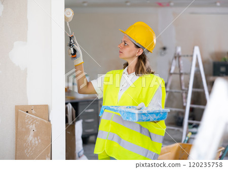 Professional female painter in protective helmet and yellow vest paints the walls with paint roller Professional female painter in protective helmet and yellow vest paints the walls with paint roller 120235758