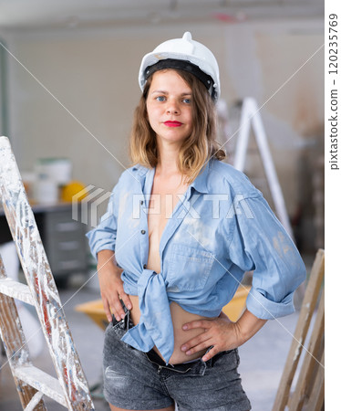 Seductive woman in hardhat and denim shirt posing next to a stepladder in room being renovated 120235769
