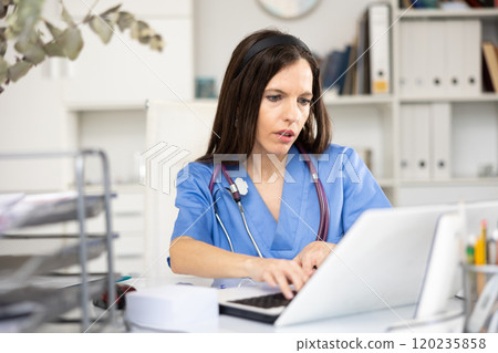Woman doctor sitting at table in her office and using laptop Woman doctor sitting at table in her office and using laptop 120235858