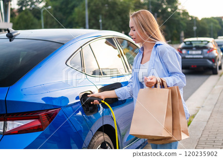 Blonde woman plugs in her electric vehicle while carrying shopping bags Blonde woman plugs in her electric vehicle while carrying shopping bags 120235902