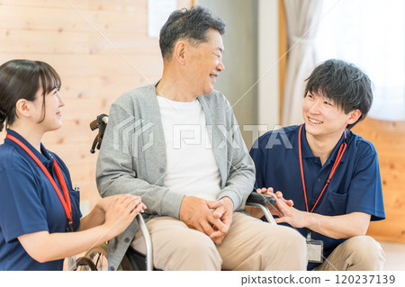 一位女性語言治療師和物理治療師微笑著與一位坐在輪椅上的老年男性患者交談（癡呆症/康復） 120237139
