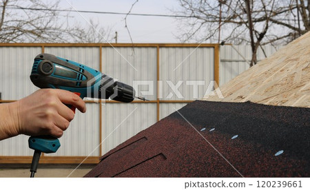 a hand with a screwdriver and a screw at the end is directed towards the roof of a low outbuilding with soft tiles to secure the material 120239661