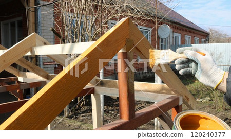 a gloved hand paints or varnishes a wooden structure with a frame made of bars against the background of a red brick house, a protective coating for wood during the construction of an outbuilding a gloved hand paints or varnishes a wooden structure with a frame made of bars against the background of a red brick house, a protective coating for wood during the construction of an outbuilding 120240377