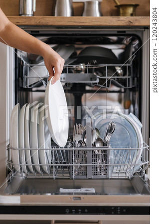 Front view from female hand removing clean plate from a loaded dishwasher. Background of modern kitchen 120243804