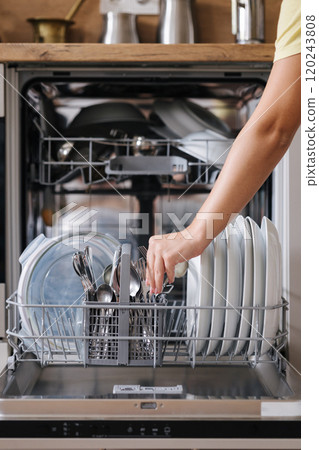 Front view from female hand removing clean cutlery from a loaded dishwasher. Background of modern kitchen 120243808