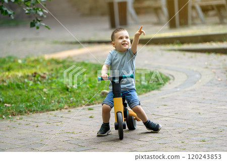 A cute smiling toddler boy of three or four years old rides a bicycle or balance bike in a city park on a sunny summer day. Toddlerhood and childhood concept. Selective focus A cute smiling toddler boy of three or four years old rides a bicycle or balance bike in a city park on a sunny summer day. Toddlerhood and childhood concept. Selective focus 120243853