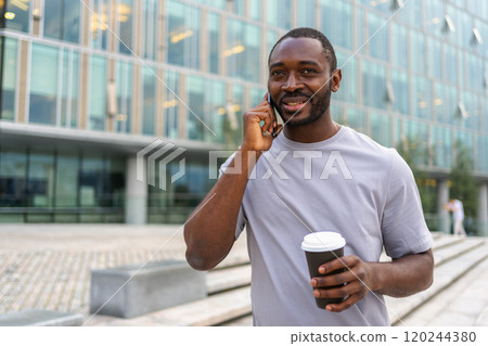 Good morning. African man drinking coffee to go talking on smartphone outdoor. Man with cell phone paper cup walking on street. Man making answering call by cellphone having conversation by mobile 120244380