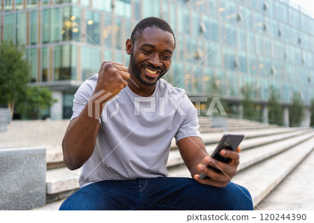 Happy African American man euphoric winner with smartphone on street in city. Person guy looking at cell phone reading great news getting good result winning online bid feeling amazed. Winning gesture Happy African American man euphoric winner with smartphone on street in city. Person guy looking at cell phone reading great news getting good result winning online bid feeling amazed. Winning gesture 120244390