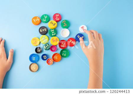 Child hands arranging colorful bottle caps with multiplication problems on blue background, flat lay educational concept for kids learning math through games, multiplication table activity 120244413