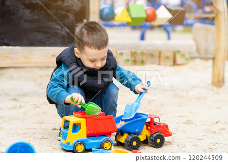 Little cute toddler boy three years old plays in the sandbox on a spring day. Outdoor development activities for kids. Toy cars and shovel 120244509
