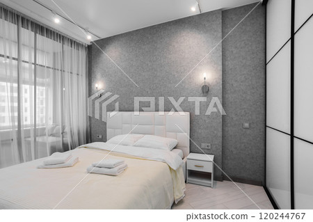 A well-appointed small modern bedroom with a cozy design, featuring a queen-sized bed, bedside tables. A large window with curtains allowing soft daylight. A well-appointed small modern bedroom with a cozy design, featuring a queen-sized bed, bedside tables. A large window with curtains allowing soft daylight. 120244767