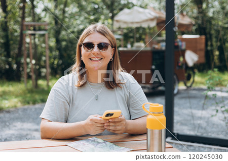 Young smiling woman in sunglasses sitting in park at cafe with smartphone and a thermos of coffee. Female explorer looking at a map outdoors, search location on phone. Lifestyle adventure concept 120245030