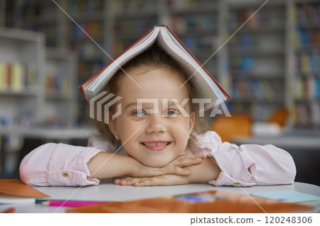 Portrait of cheerful happy girl child with book on head over library background Portrait of cheerful happy girl child with book on head over library background 120248306