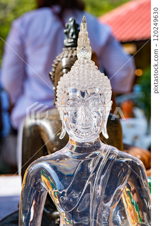 Transparent glass buddha statue in meditation posture among various buddha statues on the blurry background of people and buildings in Thailand temple. 120249630