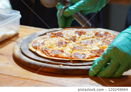 A person wearing green gloves is cutting thin-crust, small-size pizzas on a wooden tray with a pizza cutter. 120249664