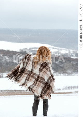 Full-length portrait of ginger hair girl wears plaid warm blanket against winter mountains. Back view. 120249782