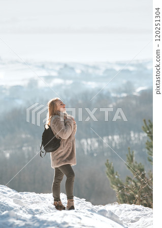 A girl in a sheepskin coat stands on a hill and looks into the distance in winter forest 120251304