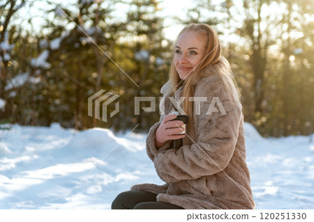 Young woman with cup of coffee sitting on snow in winter forest. 120251330