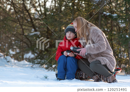 Mother and child spending time outdoors in winter. Woman pours hot tea from thermos into mug. 120251491