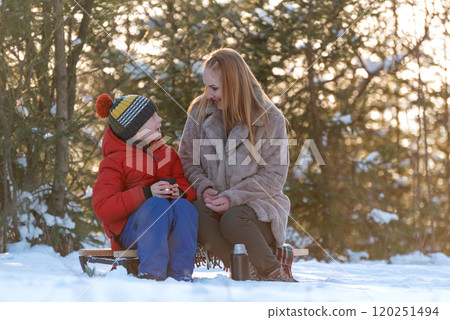 Portrait of young woman and child boy during picnic in winter in forest. Mother and son drink hot tea from thermos. Portrait of young woman and child boy during picnic in winter in forest. Mother and son drink hot tea from thermos. 120251494