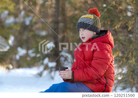 Boy in red jacket and hat sitting on the snow with a cup in his hand, sits and looks into the distance Boy in red jacket and hat sitting on the snow with a cup in his hand, sits and looks into the distance 120251500