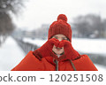Portrait of girl in red winter jacket and woolen hat with mittens showing heart with hands. Woman in fashionable winter look outdoors 120251508
