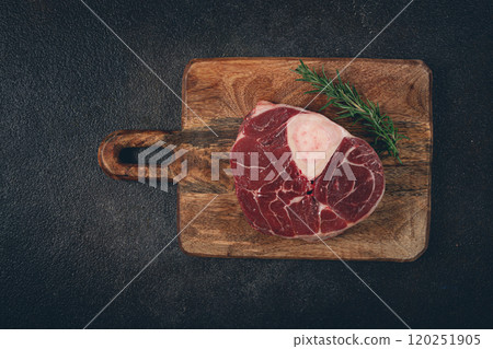 a shank on the bone, raw meat for ossobuco, on a wooden chopping board, with a branch of rosemary, top view, no people. 120251905
