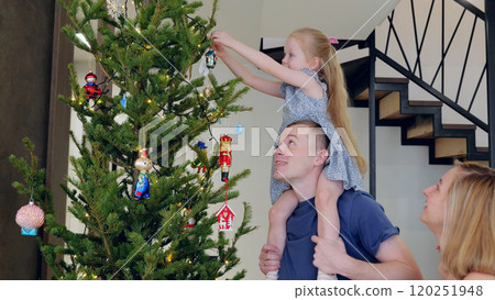 Joyful family decorates a grand christmas tree in their cozy living room, embracing the festive spirit of the holiday season. Daughter decorates a Christmas tree while sitting on her dad's neck 120251948