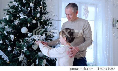 Father and son happily decorate a christmas tree with silver and white ornaments in a cozy living room, creating a festive atmosphere of love and togetherness. A day with family before Christmas.  120251962
