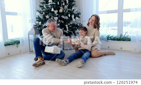 Happy family gathers around a christmas tree, sharing gifts in a cozy living room filled with festive decorations and warm light. A day with family before Christmas. New Year's Eve with family 120251974