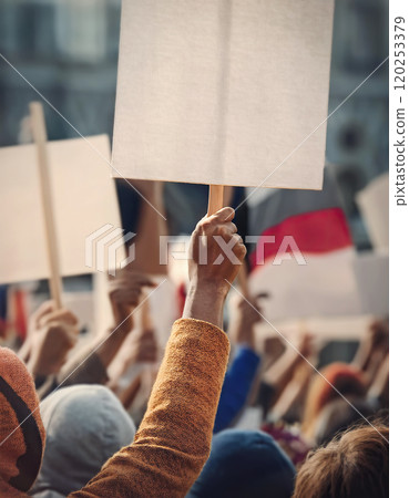 Crowd of people on the street holding up blank protest banners. Hooded man in the foreground holding a placard. Scene of demonstration, riot or rally 120253379