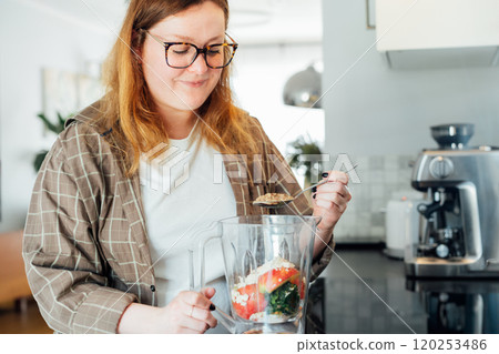 Plus size young woman adding flex seeds during making smoothie on the kitchen. Superfood supplement. Healthy detox vegan diet. Healthy dieting eating, weight loss program. Selective focus. Plus size young woman adding flex seeds during making smoothie on the kitchen. Superfood supplement. Healthy detox vegan diet. Healthy dieting eating, weight loss program. Selective focus. 120253486