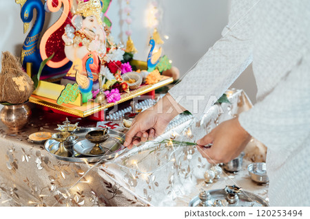 Close up Indian man dressed in traditional wear engages in worship at home altar, adorned with decors and offerings for god Ganesh during festive celebration. Indian culture, hindu ritual and customs 120253494