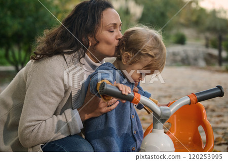 Mother kissing toddler on tricycle in a scenic park 120253995