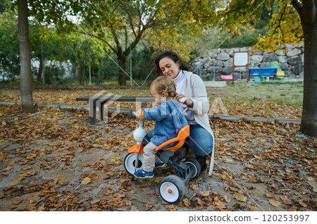 Mother guiding toddler on tricycle in autumn park scene Mother guiding toddler on tricycle in autumn park scene 120253997
