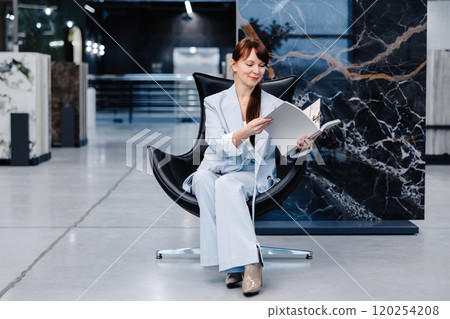 Woman in a light blue suit sitting in a black chair holding a white book in a modern showroom Woman in a light blue suit sitting in a black chair holding a white book in a modern showroom 120254208