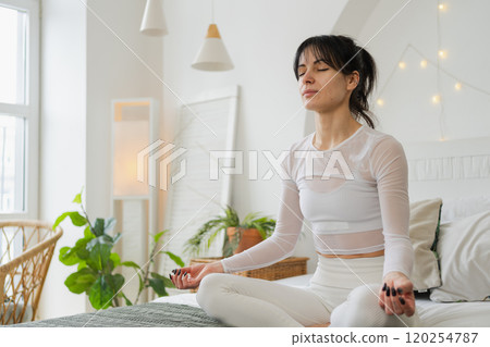 Yoga mindfulness meditation. Young healthy woman practicing yoga in bedroom at home. Woman sitting in lotus pose on bed meditating smiling relaxing indoor. Girl doing breathing practice. Yoga at home Yoga mindfulness meditation. Young healthy woman practicing yoga in bedroom at home. Woman sitting in lotus pose on bed meditating smiling relaxing indoor. Girl doing breathing practice. Yoga at home 120254787
