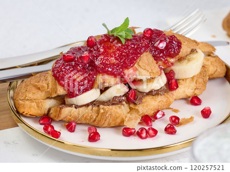 Croissant with banana pieces, chocolate paste and pomegranate seeds in a round white plate Croissant with banana pieces, chocolate paste and pomegranate seeds in a round white plate 120257521