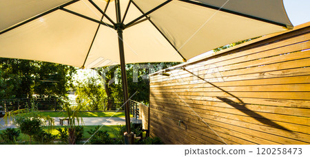 Sunlit outdoor umbrella and wooden fence with natural greenery. Concept of shade, privacy, and peaceful outdoor retreat 120258473