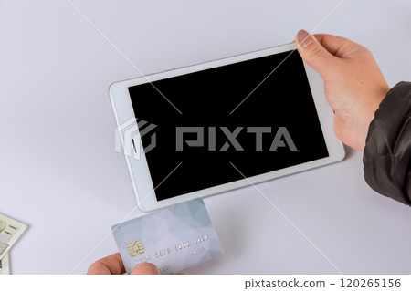 Person holds credit card near tablet while preparing to make an online purchase surface, suggesting financial transactions. Person holds credit card near tablet while preparing to make an online purchase surface, suggesting financial transactions. 120265156
