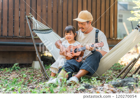 Father and daughter playing ukulele while sitting in a hammock at a campsite, outdoor leisure Father and daughter playing ukulele while sitting in a hammock at a campsite, outdoor leisure 120270054