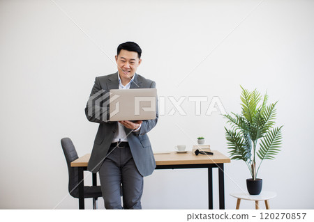 Smiling businessman working on laptop in modern office space 120270857