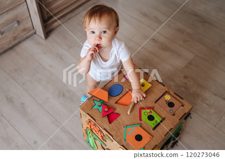 Happy toddler baby boy is playing with a busyboard cube on the nursery floor. A child aged one year Happy toddler baby boy is playing with a busyboard cube on the nursery floor. A child aged one year 120273046