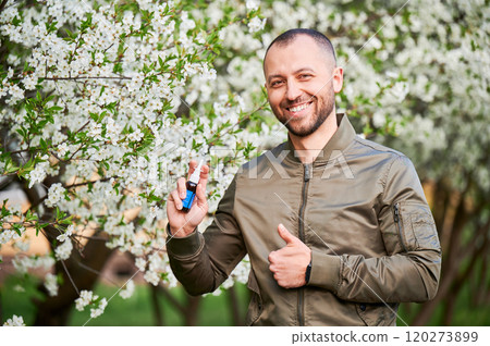 Man allergic using medical nasal spray, suffering from seasonal allergy at spring in blossoming garden. Portrait of smiling man showing thumbs up near blooming tree outdoors. Spring allergy concept. Man allergic using medical nasal spray, suffering from seasonal allergy at spring in blossoming garden. Portrait of smiling man showing thumbs up near blooming tree outdoors. Spring allergy concept. 120273899