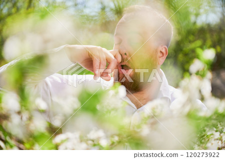 Man allergic suffering from seasonal allergy at spring in blossoming garden at springtime. Close up of bearded man sneezing in front of blooming tree. Spring allergy concept. 120273922