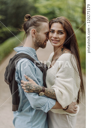 A beautiful couple's engagement portrait by the lovely trees. A stylish young couple, dressed in elegant attire, embraces each other in a lush green field, expressing love and connection. Warsaw. 120275179
