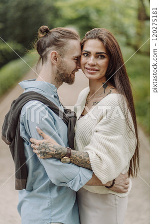 A beautiful couple's engagement portrait by the lovely trees. A stylish young couple, dressed in elegant attire, embraces each other in a lush green field, expressing love and connection. Warsaw. 120275181