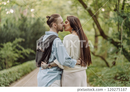 A beautiful couple's engagement portrait by the lovely trees. A stylish young couple, dressed in elegant attire, embraces each other in a lush green field, expressing love and connection. Warsaw. 120275187