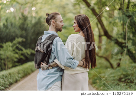 A beautiful couple's engagement portrait by the lovely trees. A stylish young couple, dressed in elegant attire, embraces each other in a lush green field, expressing love and connection. Warsaw. 120275189